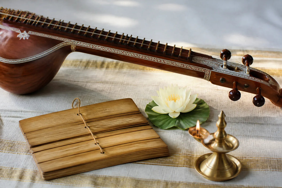 A stringed instrument, blank palm-leaf-style manuscript, white lotus, and lit brass lamp arranged on pale cloth.