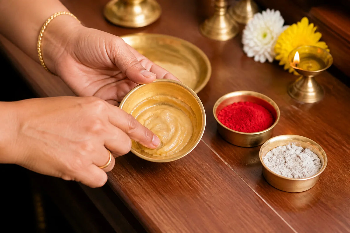Hands preparing a pale paste in a brass bowl beside red and white powders, flowers, and a small lit brass lamp.
